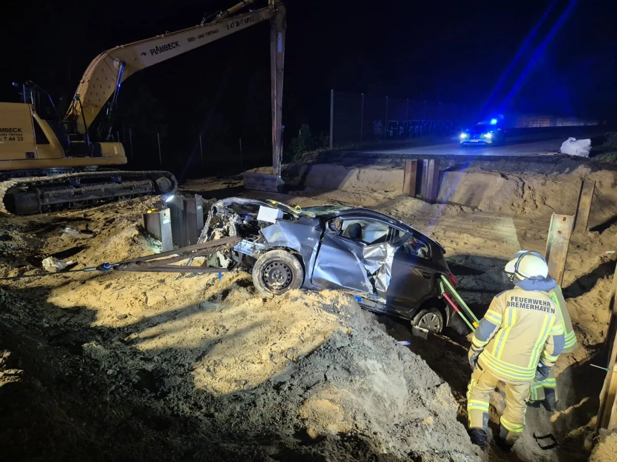 Ein beschädigtes Auto liegt in einer Baugrube, während ein Feuerwehrmann in voller Ausrüstung danebensteht. Ein Bagger ist im Hintergrund zu sehen.