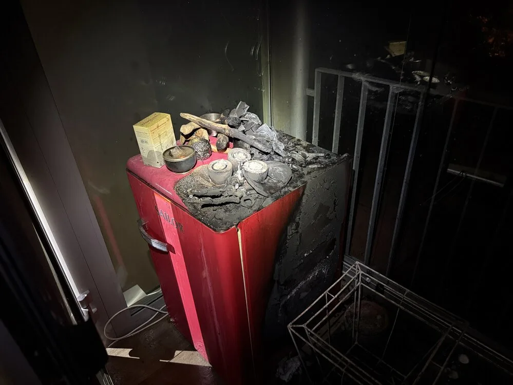 Ein roter Kühlschrank auf einem Balkon zeigt deutliche Brandschäden. Auf ihm befinden sich verkohlte Gegenstände, die auf ein Feuer hinweisen.