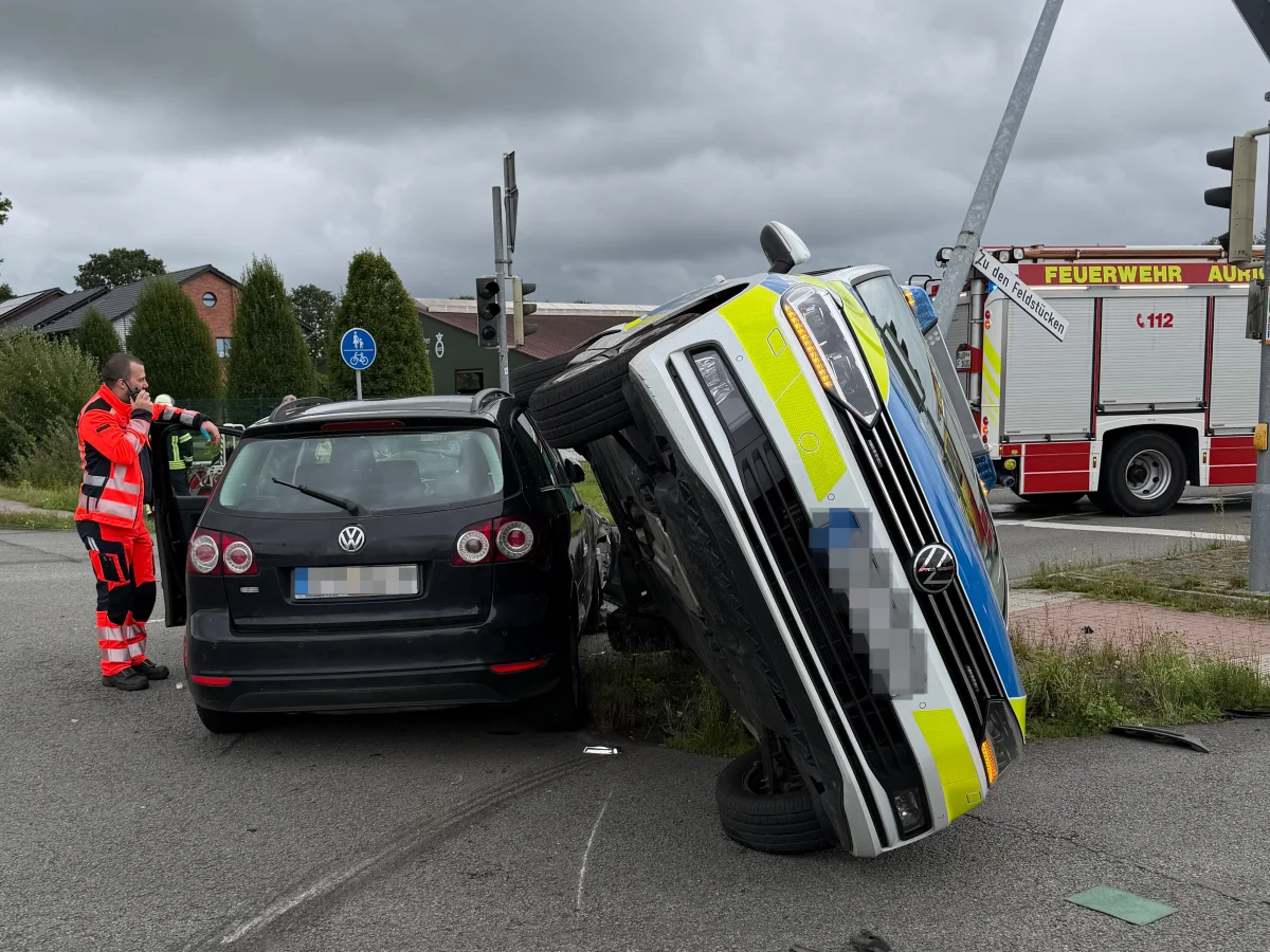 Ein umgekippter Polizeiwagen liegt auf der rechten Seite neben einem schwarzen VW Golf. Ein Feuerwehrfahrzeug ist im Hintergrund zu sehen.