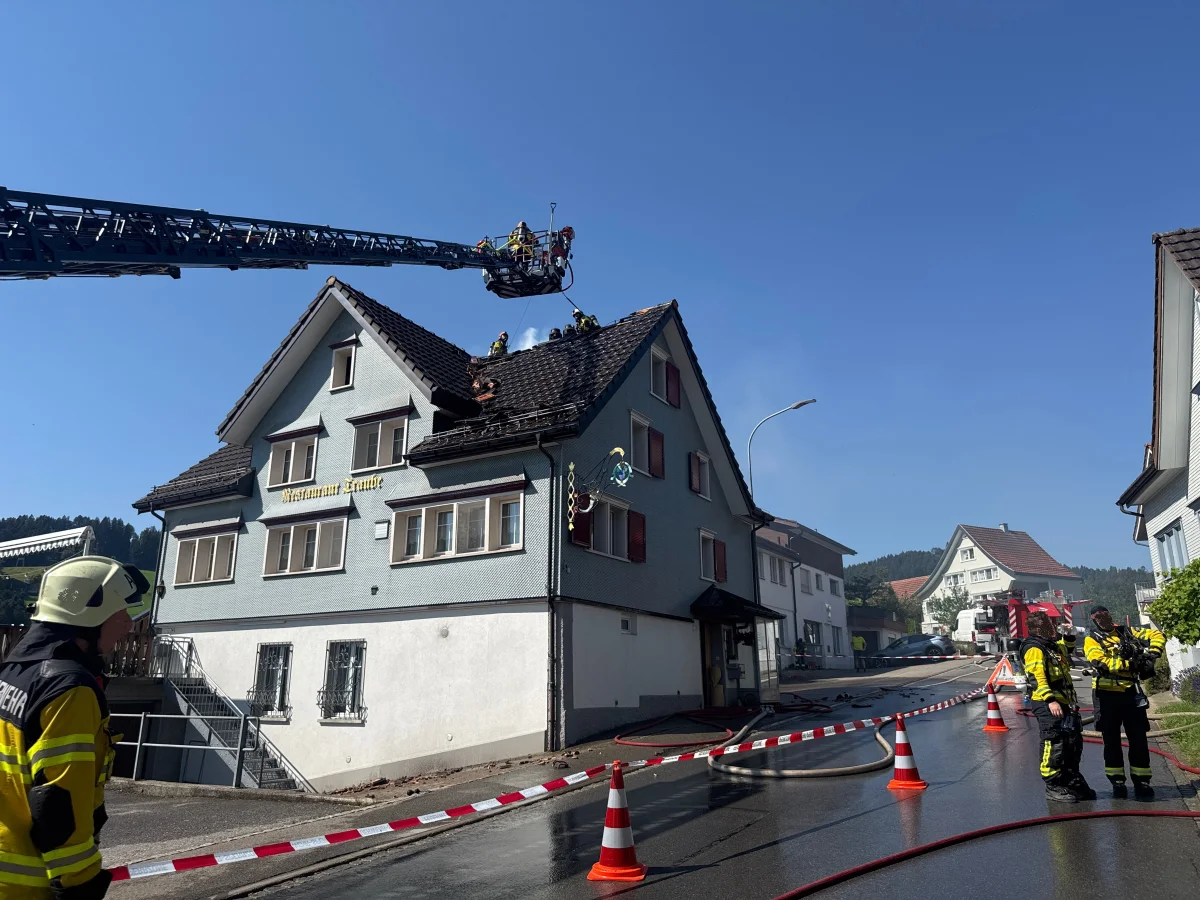 Mehrere Feuerwehrleute sind auf dem Dach eines Gebäudes mit einer Drehleiter in Aktion. Der Himmel ist klar und blau.