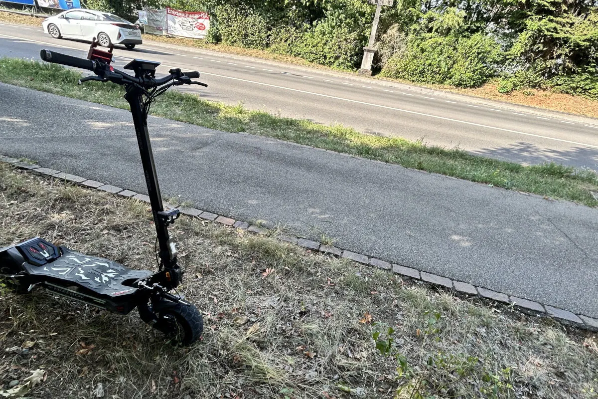 Ein schwarzer E-Scooter steht am Rand eines mit Bäumen gesäumten Gehweges. Im Hintergrund ist eine Straße zu sehen.