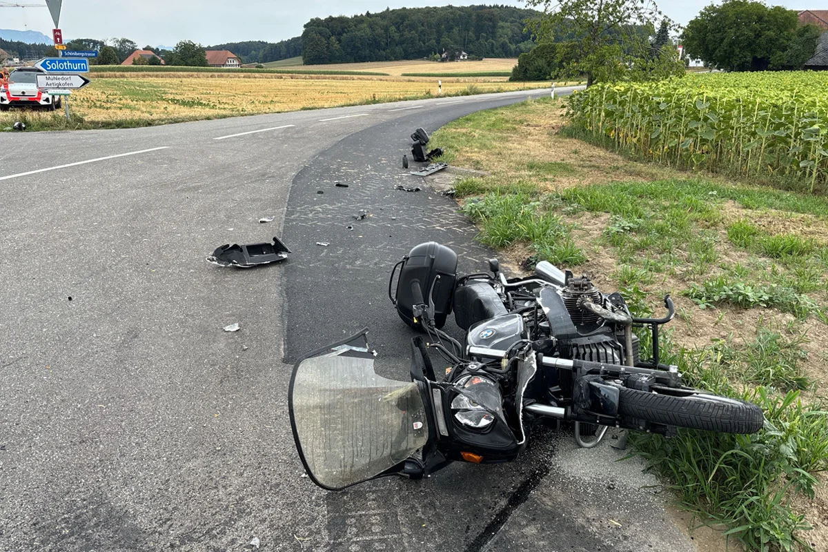 Ein schwarzes Motorrad liegt auf der Seite neben Trümmern auf einer Landstraße, umgeben von Feldern und einem Verkehrszeichen Richtung Solothurn.