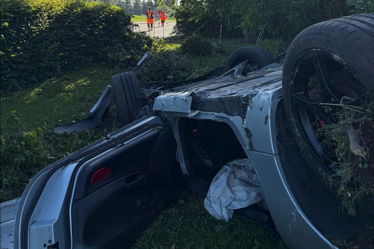 Ein silbernes Auto liegt auf dem Dach im Gras neben einer Hecke. Der Innenraum des Autos ist sichtbar. Im Hintergrund stehen Personen in orangefarbenen Westen.