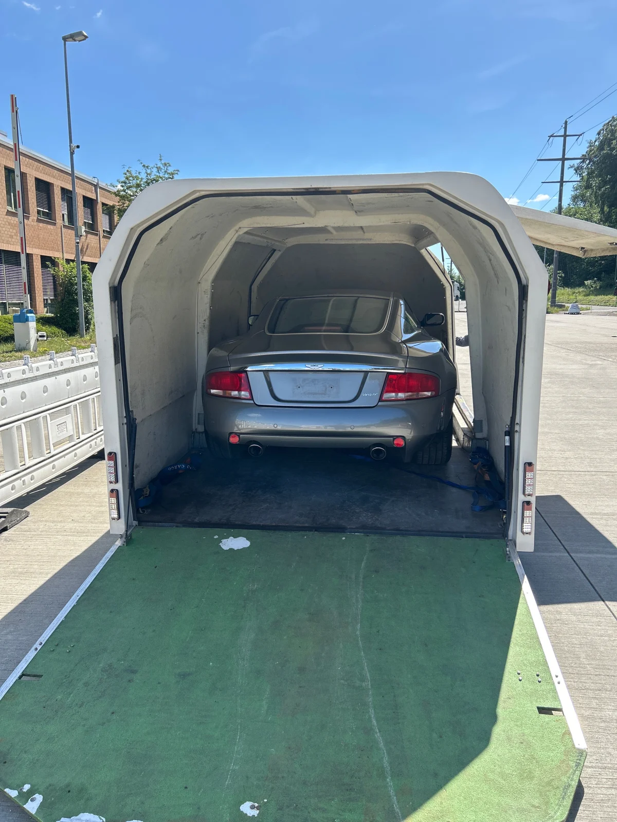 Ein silberfarbenes Auto steht in einem geöffneten Anhänger. Das Bild zeigt eine sonnige Umgebung mit klarem Himmel und wenigen sichtbaren Personen oder Fahrzeugen außer dem Anhänger.