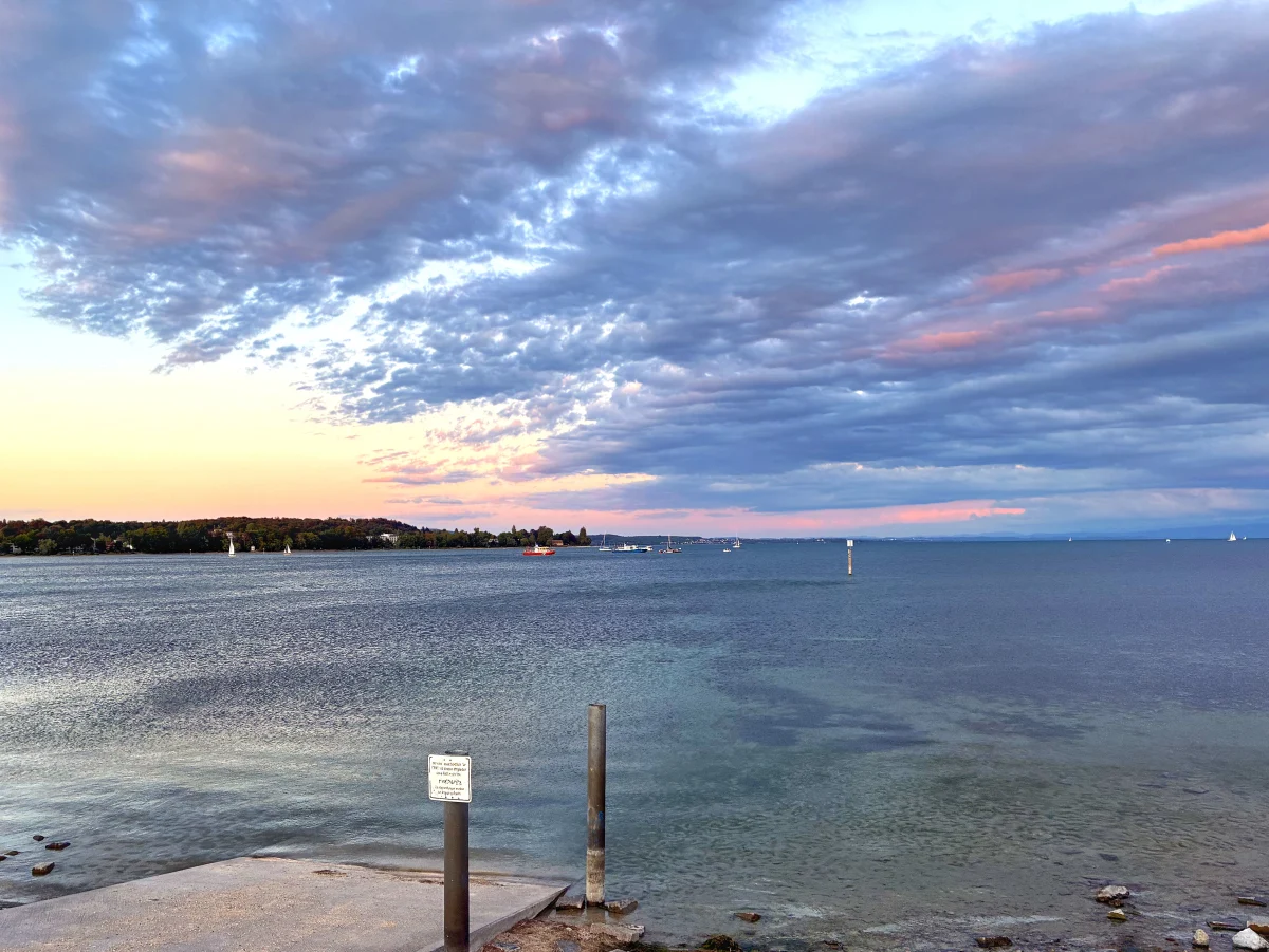 Der See zeigt ein einzelnes Boot bei bewölktem Himmel während der Dämmerung. Der Horizont ist mit Bäumen gesäumt.