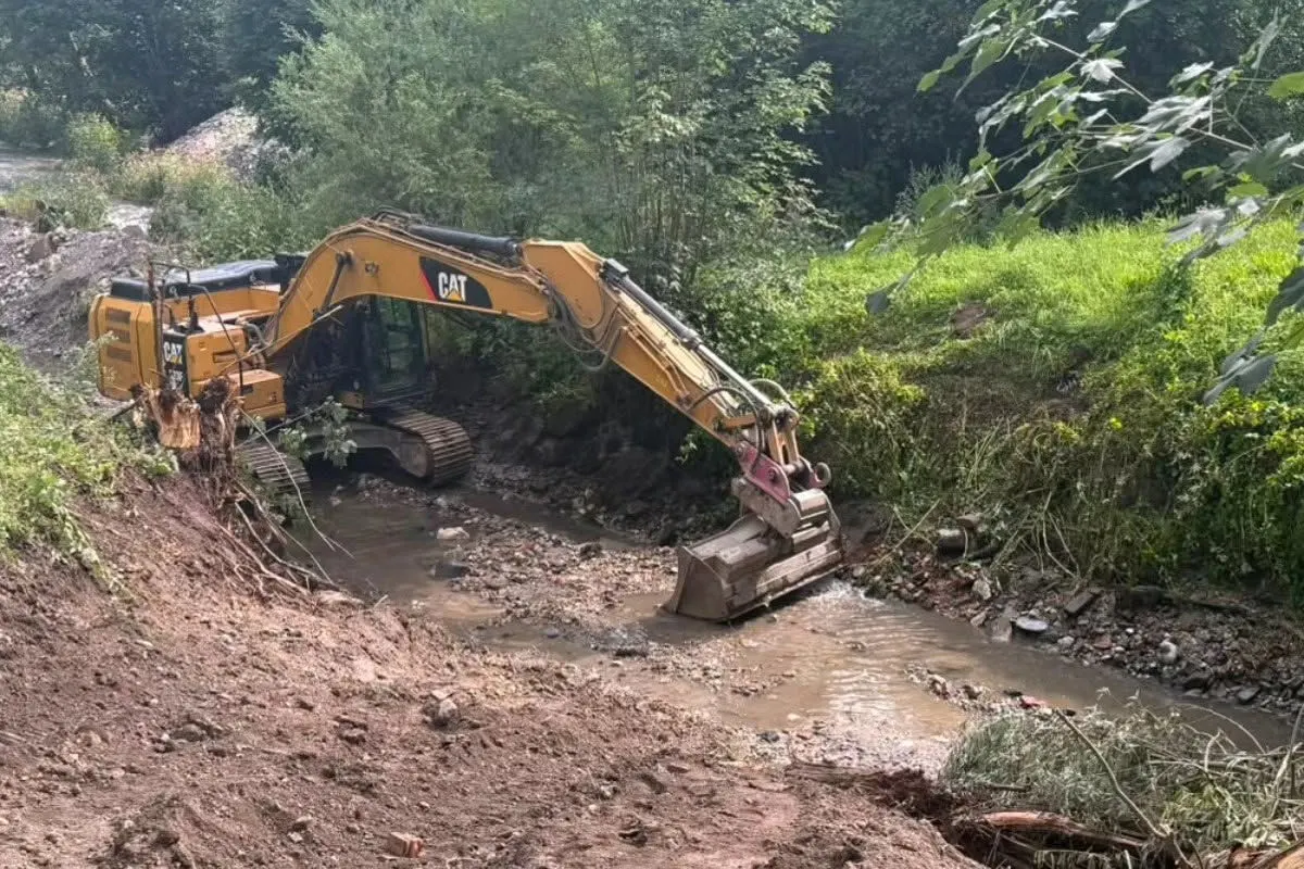 Ein gelber Bagger ist in einem Bachbett in einem waldreichen Gebiet im Einsatz. Der Boden um den Bach ist uneben und mit Erde bedeckt.