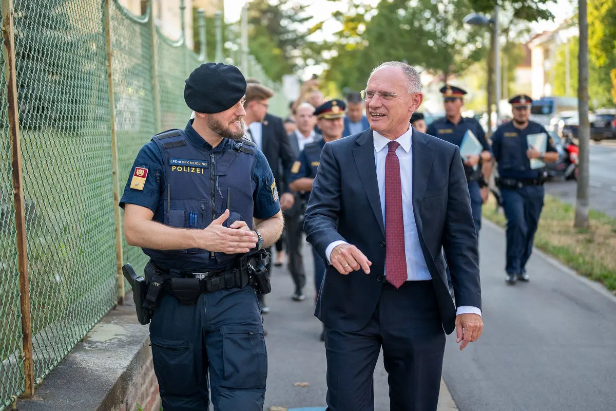Ein Polizist in Uniform unterhält sich mit einem Mann im Anzug auf einem Gehweg. Im Hintergrund sind weitere Personen zu sehen.