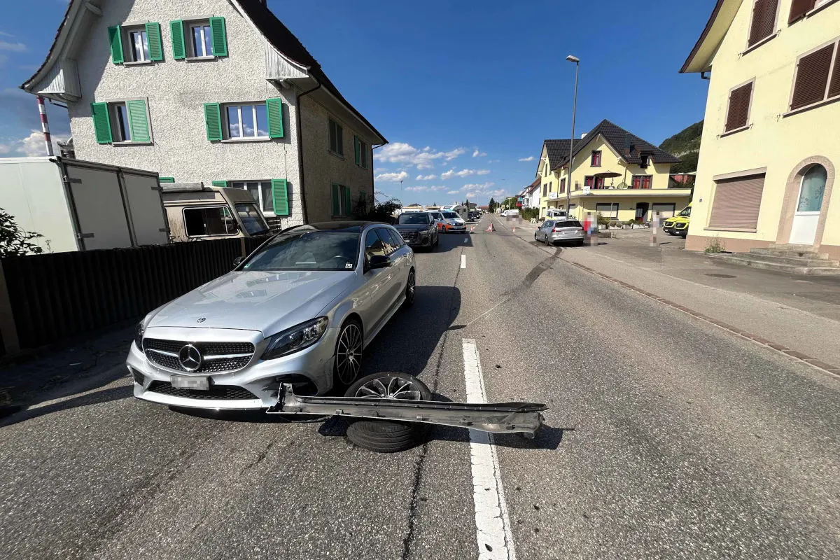 Ein silberner Mercedes mit abgerissenem Stoßfänger steht auf einer Straße, umgeben von Wohnhäusern.