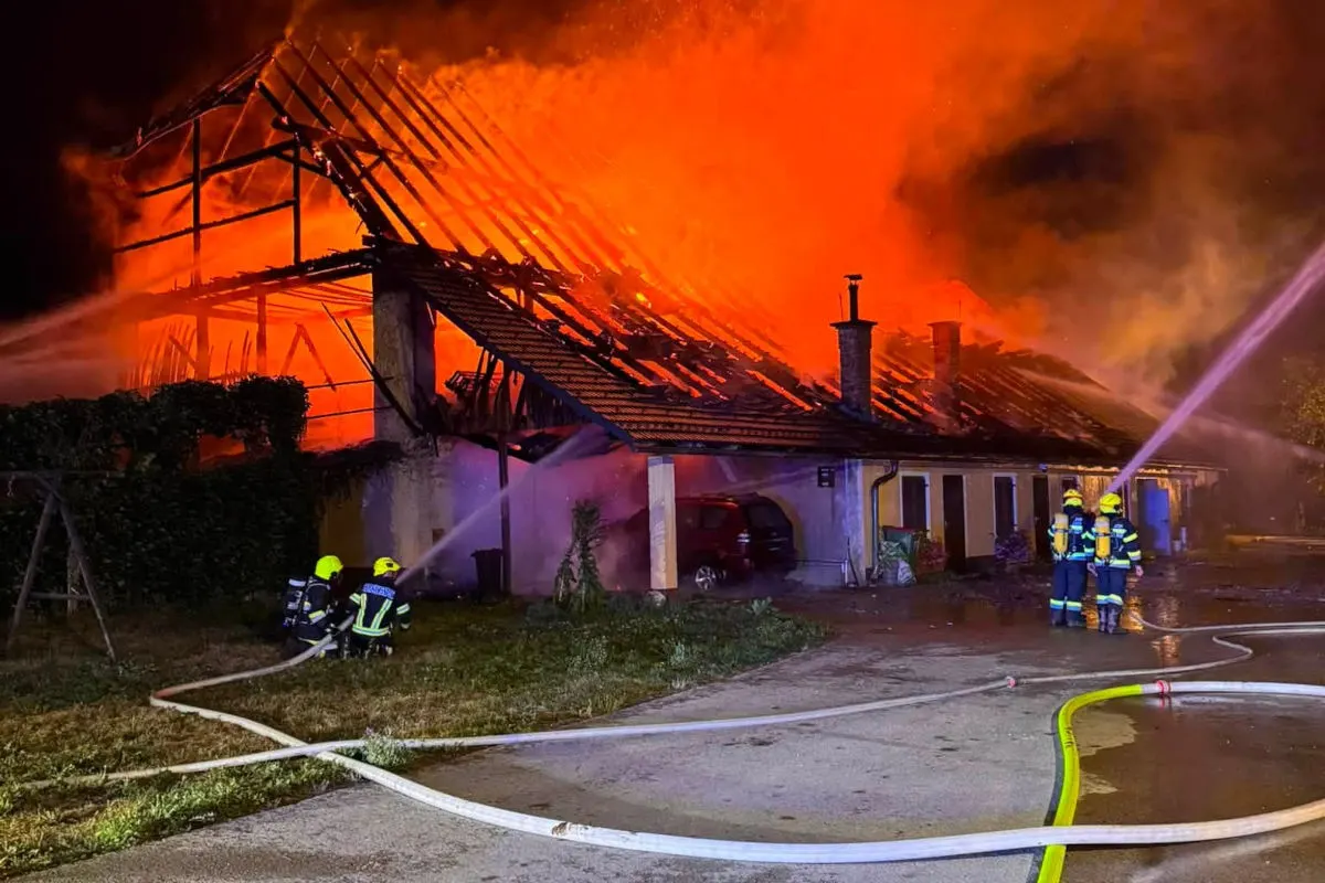 Mehrere Feuerwehrleute bekämpfen in der Nacht einen intensiven Brand in einem großen Wohnhaus. Flammen schlagen aus dem Dachstuhl, während Wasser aus Schläuchen auf das Feuer gerichtet wird.