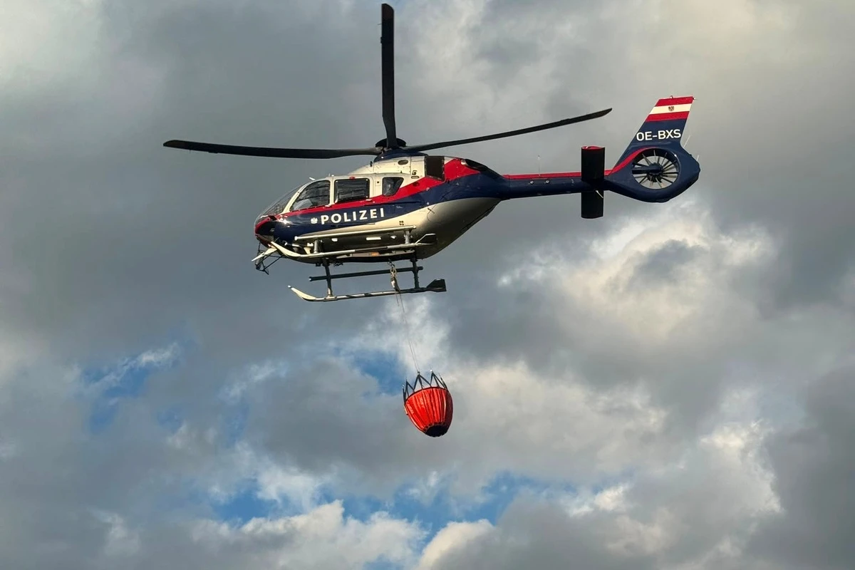 Ein Polizeihubschrauber fliegt am Himmel mit einem roten Löschwasserbehälter. Der Hintergrund ist von Wolken bedeckt.