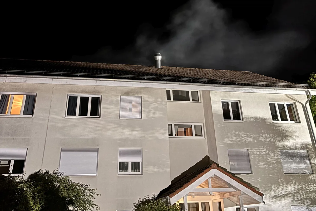 Ein Wohngebäude bei Nacht, aus dessen Schornstein Rauch aufsteigt. Mehrere Fenster sind teilweise beleuchtet.