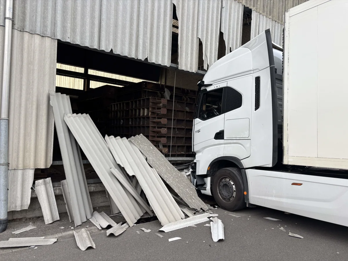 Ein weißer Lkw ist in die Wand einer Lagerhalle gefahren, wobei mehrere Wellblechplatten beschädigt wurden. Die Halle zeigt deutliche strukturelle Schäden.