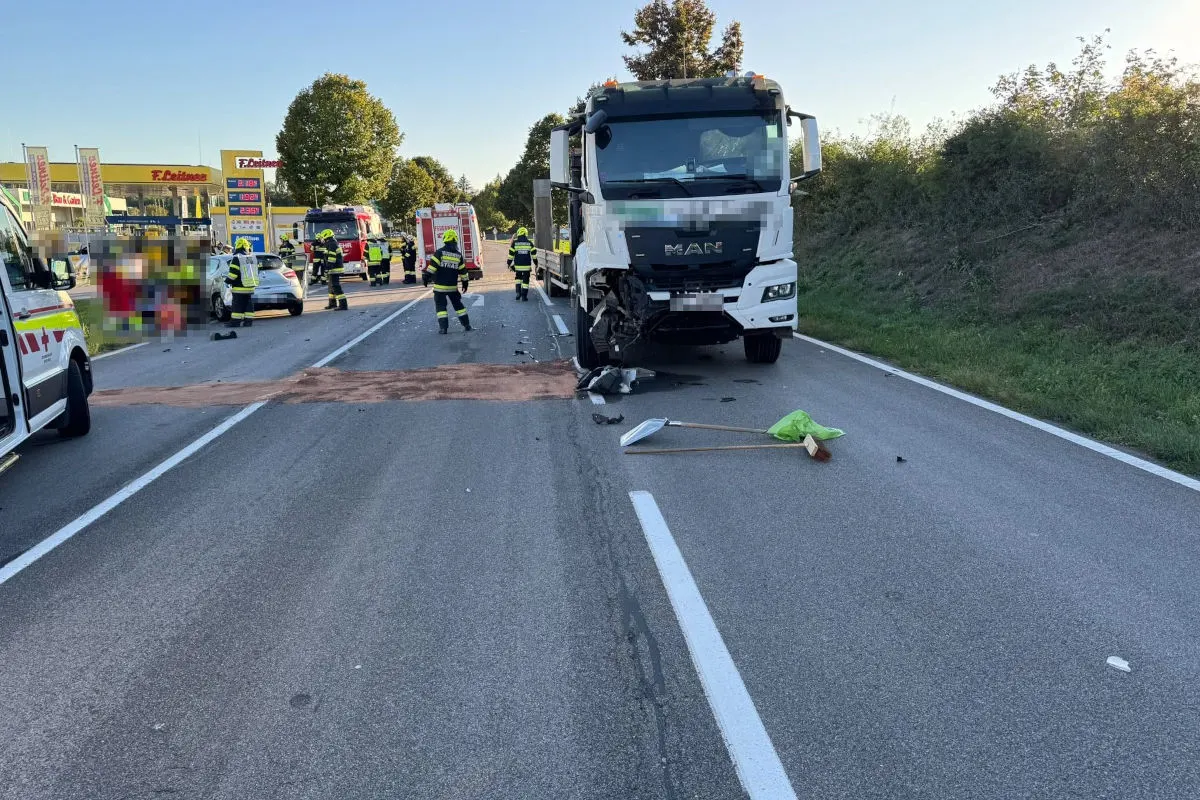 Ein weißer Lastwagen mit deutlichen Schäden steht auf einer Straße. Feuerwehrleute und Rettungsfahrzeuge sind im Hintergrund sichtbar.