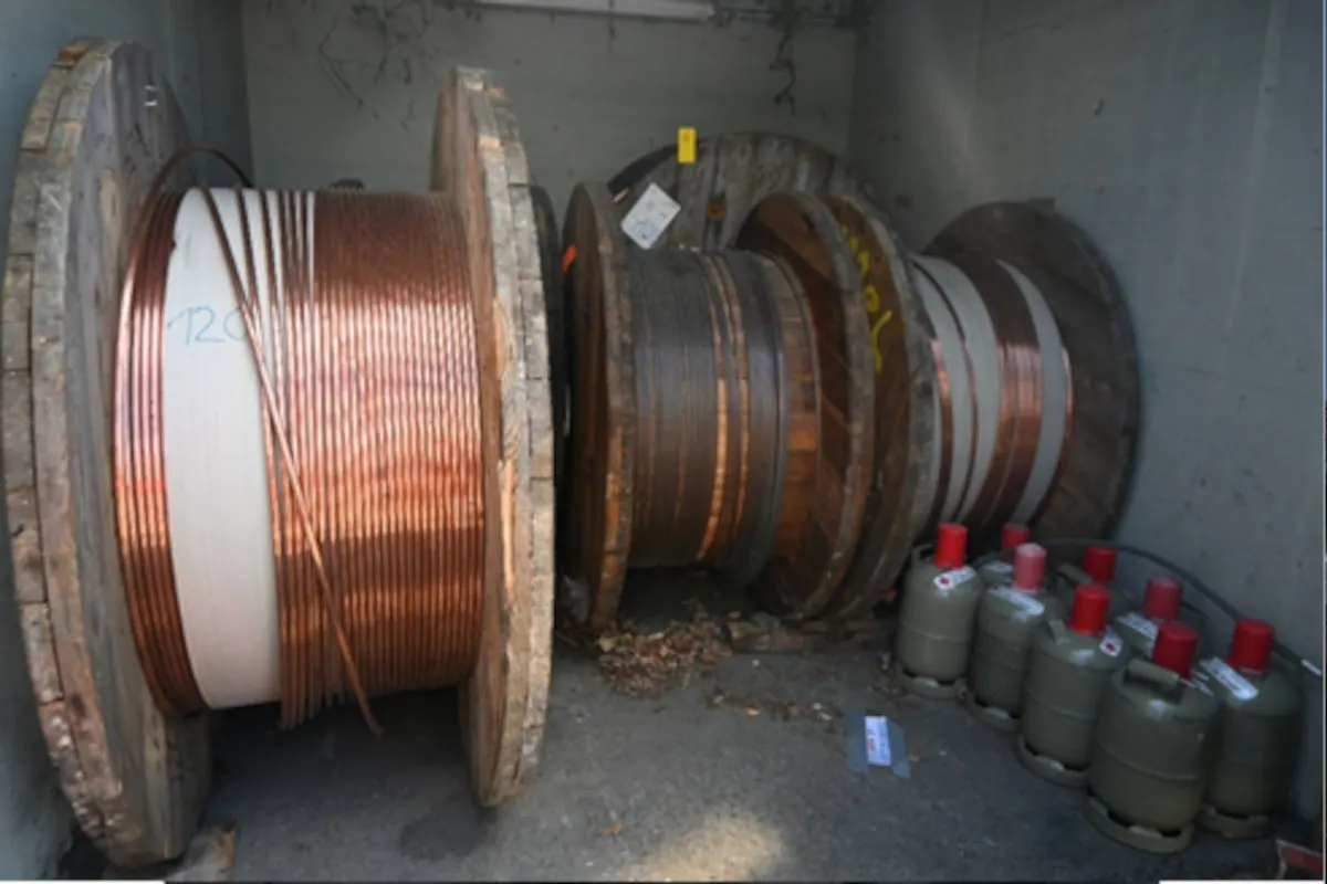 Mehrere Kabeltrommeln aus Holz und Metall sowie Gasflaschen sind in einem kleinen Raum gelagert. Der Boden ist mit Laub bedeckt, und die Umgebung wirkt ungepflegt.