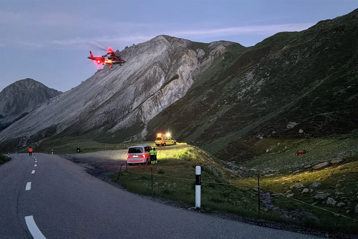 Ein Rettungshubschrauber schwebt bei Dämmerung über einer kurvigen Bergstraße. Ein Einsatzfahrzeug ist am Straßenrand zu sehen.