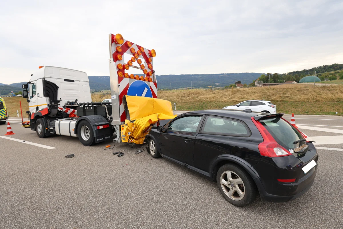 Ein schwarzer Pkw ist auf der Autobahn mit einem gelben Absicherungsanhänger zusammengestoßen. Das Fahrzeug steht direkt hinter dem Anhänger, der sichtbar beschädigt ist.