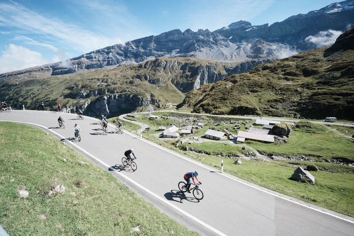 Mehrere Radfahrer fahren eine kurvige Straße in einer malerischen Alpenlandschaft hinunter. Im Hintergrund erstrecken sich imposante Berge und blauem Himmel.