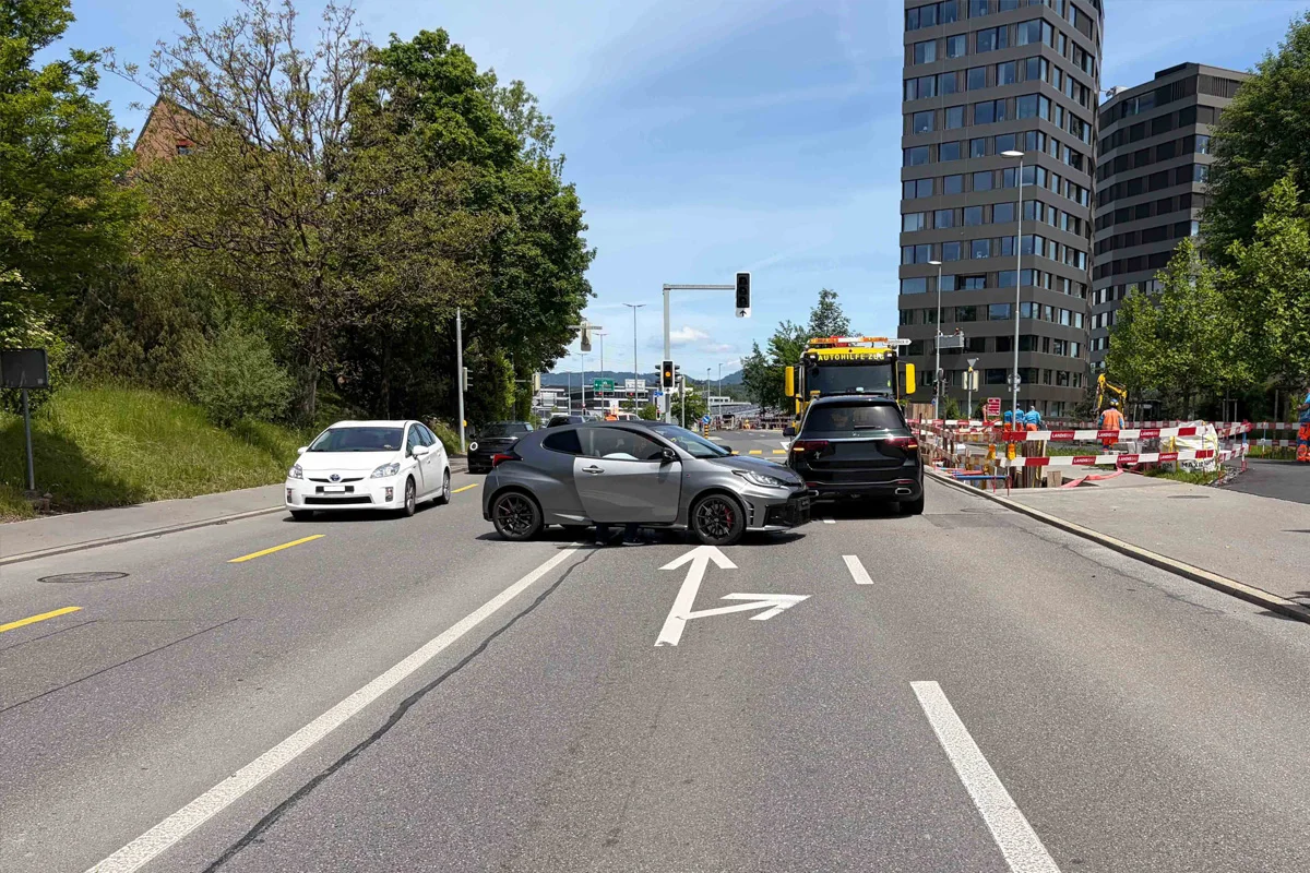 Ein grauer und ein schwarzer SUV stehen mitten auf einer vielspurigen Straße, umgeben von einem weißen PKW. Im Hintergrund ist eine Baustelle sichtbar.