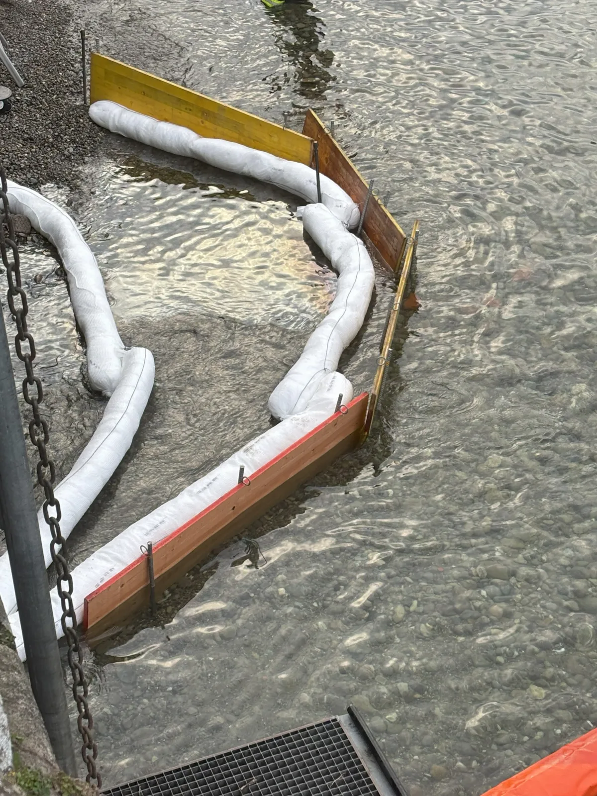 Mehrere Ölsperren sind im flachen Wasser eines Flusses platziert, um Verschmutzungen einzudämmen. Holzbarrieren und weiße Schläuche bilden eine Barriere im Wasser.