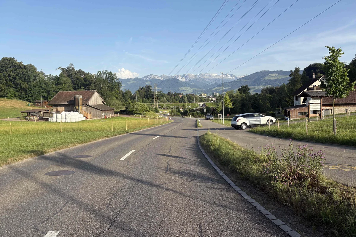 Das Bild zeigt eine ländliche Straße mit einem geparkten Auto auf der rechten Seite. Im Hintergrund ist ein Bauernhof und schneebedeckte Alpen unter klarem Himmel zu sehen.