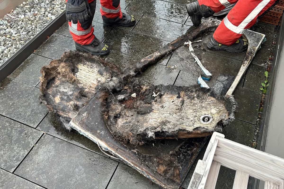 Zwei Feuerwehrleute in rot-weißen Anzügen stehen auf einer nassen, gepflasterten Fläche und untersuchen ein verkohltes Stück Material.