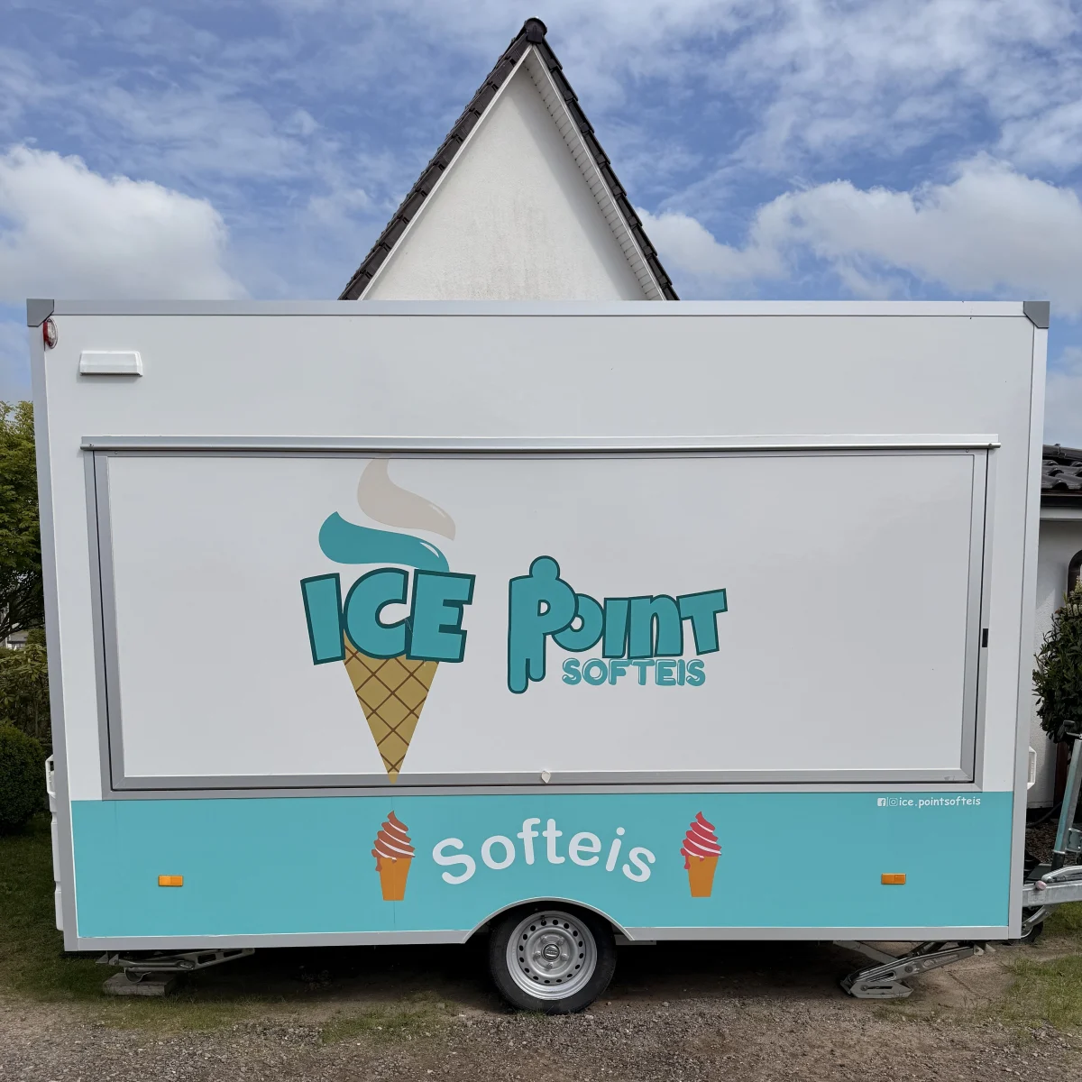Ein moderner Eiswagen mit der Aufschrift 'Ice Point Softeis' steht vor einem Gebäude mit deutlichem Giebeldach. Der Himmel ist teils bewölkt.