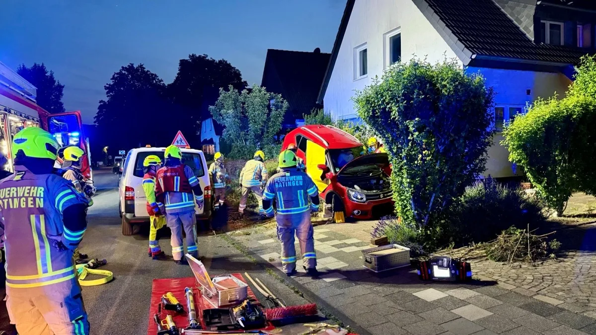 Mehrere Feuerwehrleute in Schutzkleidung sichern ein rotes Auto, das neben einem Wohnhaus beschädigt ist. Ausrüstung und Fahrzeuge sind zu sehen.