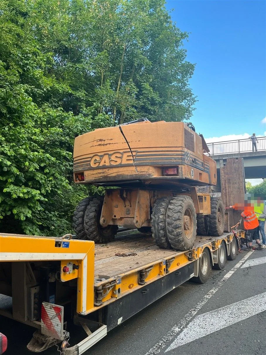Ein orangefarbener Bagger sitzt auf einem großen Transportanhänger, der sich auf einer Autobahn befindet. Ein Arbeiter, in Warnkleidung, befindet sich daneben.