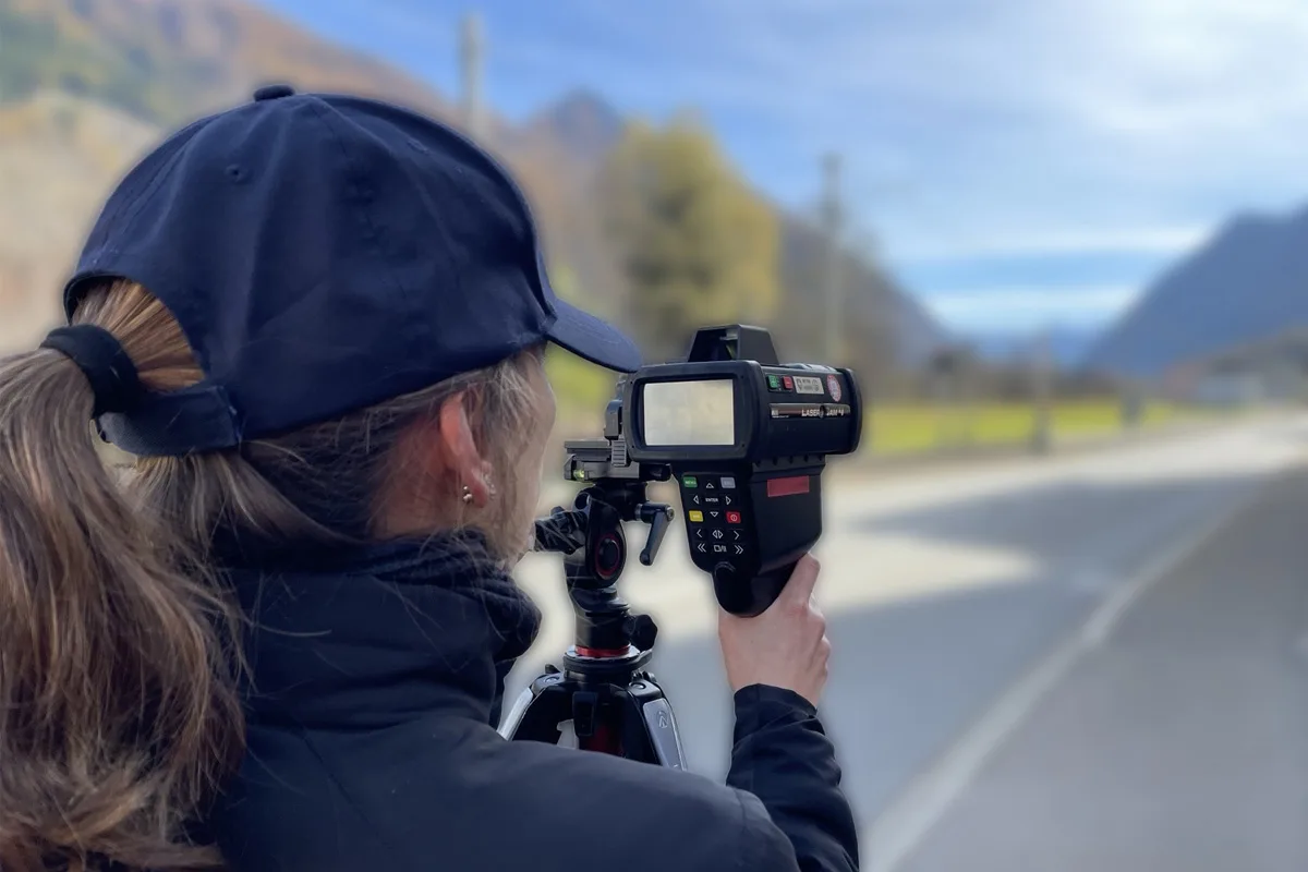 Eine Frau im Dunkelblauen Outfit verwendet ein Handradar zur Geschwindigkeitsmessung an einer Landstraße in einem ländlichen Gebiet.
