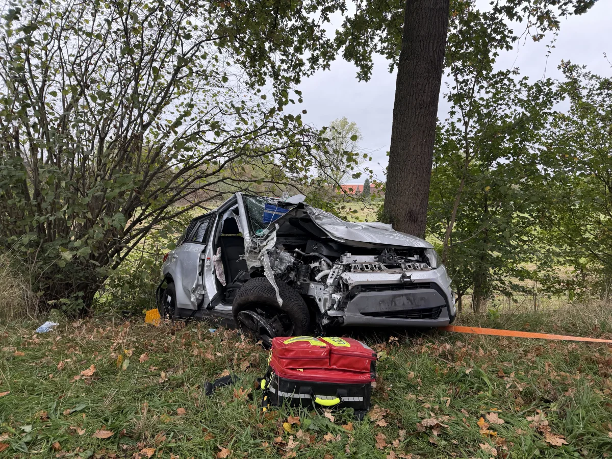 Ein stark deformiertes Auto steht nach einem Unfall gegen einen Baum. Auf der Wiese ist Rettungsgerät sichtbar.