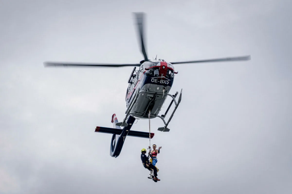Ein Hubschrauber der Rettungskräfte schwebt in der Luft, wobei zwei Personen an einem Seil unter ihm herabgelassen werden.