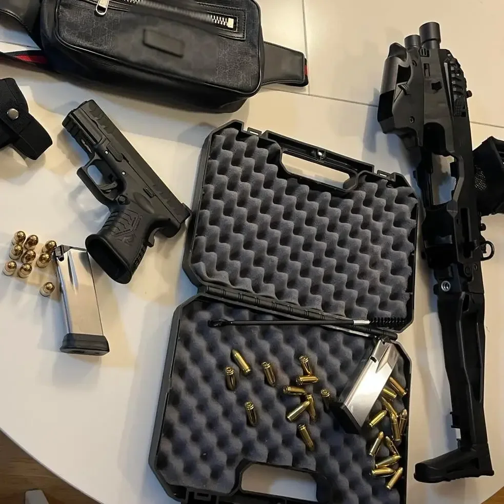 Auf einem Tisch liegen eine Pistole, ein Gewehr, Munition und eine Tasche. Ein Waffenkoffer ist geöffnet.