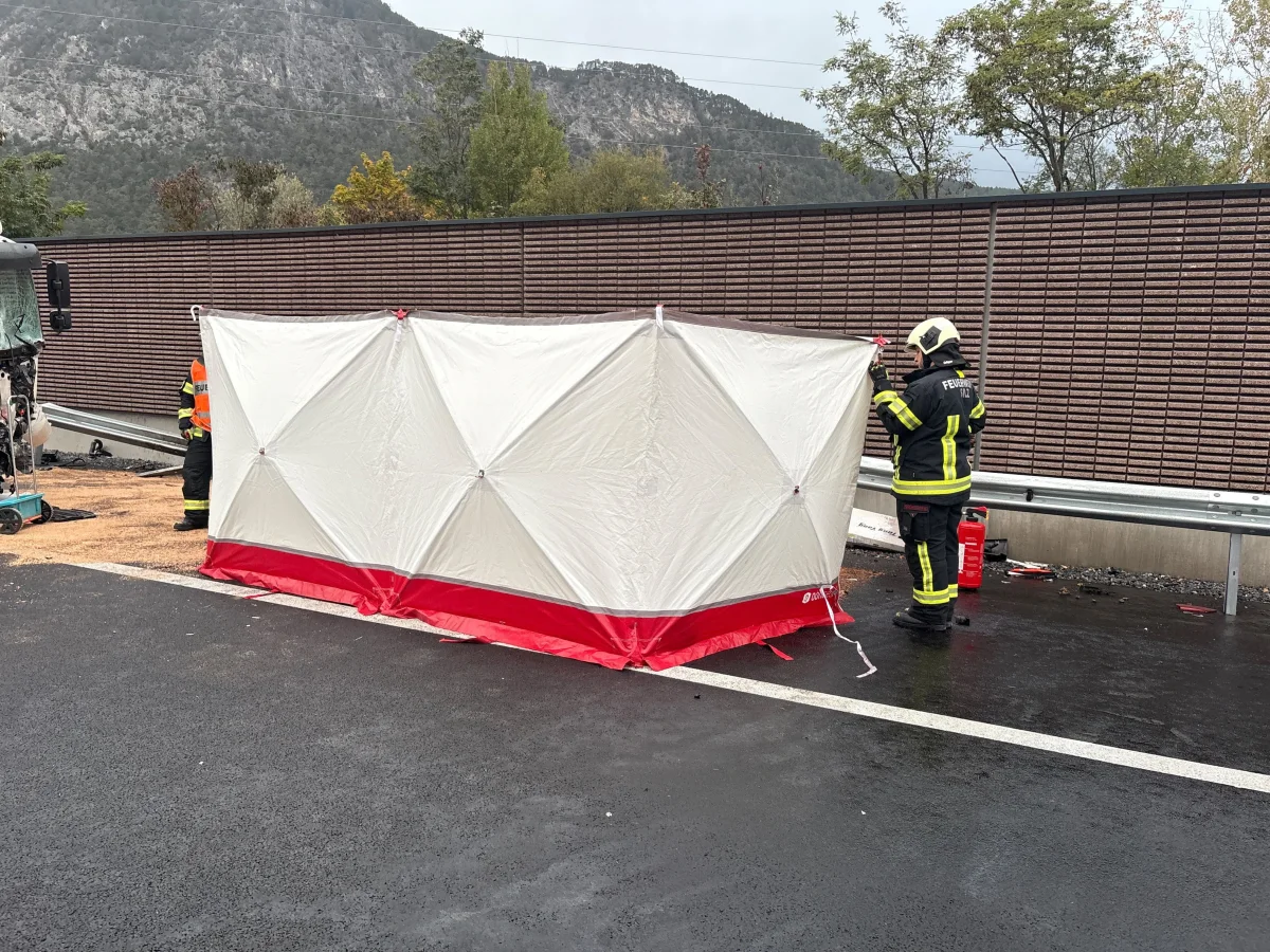 Zwei Feuerwehrleute bauen eine rot-weiße Abschirmung auf einer nassen Autobahn neben einem LKW auf. Im Hintergrund sind Bäume und eine Lärmschutzwand sichtbar.
