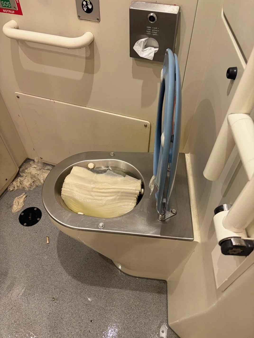Ein Zug-WC ist durch eine hineingestopfte Papierrolle verstopft. Der Boden ist unordentlich mit Abfall bedeckt.