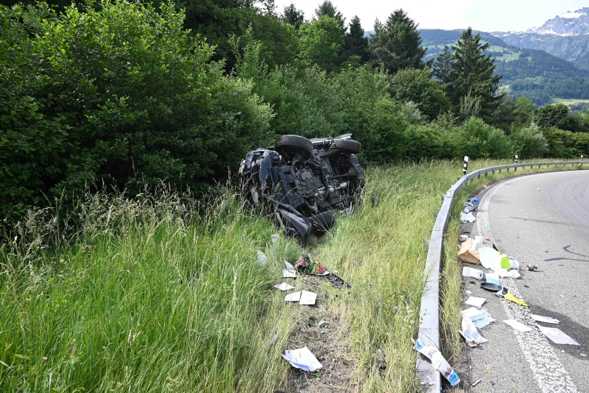 Ein Auto liegt umgestürzt auf der Seite neben einer kurvenreichen Landstraße. Rund um das Fahrzeug ist Gras und Gebüsch zu sehen.