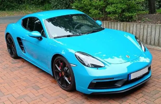 Ein hellblauer Porsche steht auf einem Backsteinpflasterparkplatz, umgeben von grüner Vegetation und einer Mauer rechts.