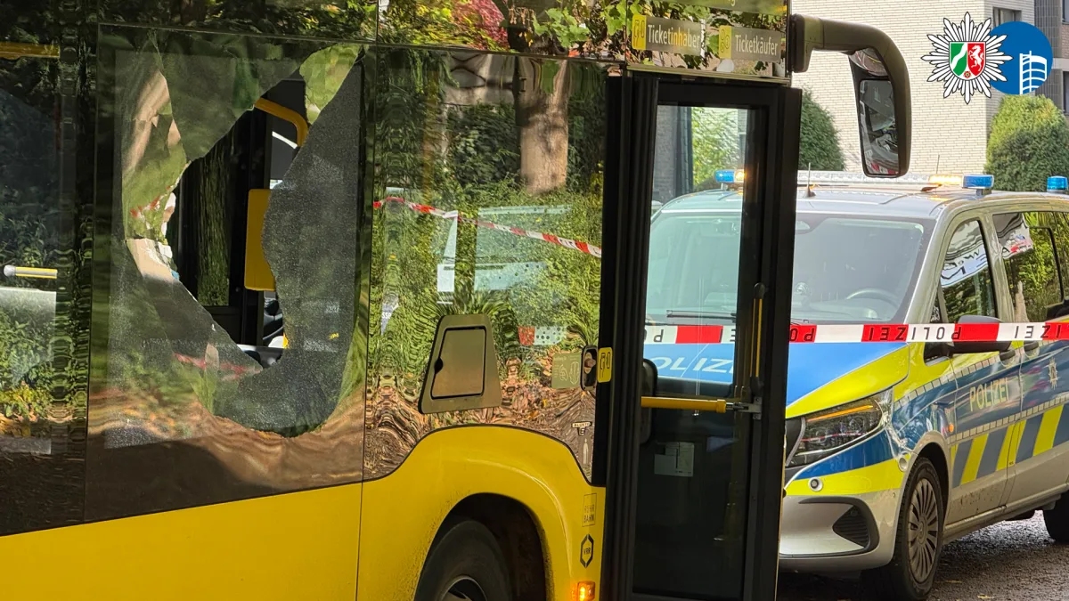 Ein Bus mit einem zerstörten Fenster, dahinter steht ein Polizeiauto mit Absperrband. Die Szene wirkt urban.