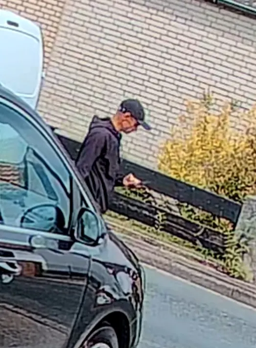 Eine Person in dunkler Kleidung und Mütze geht neben einem geparkten Auto auf einem Gehweg entlang.