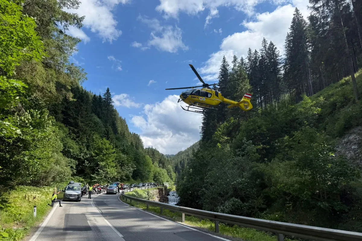 Ein gelber Rettungshubschrauber fliegt über eine schmale, kurvige Straße, die durch einen dichten Wald führt. Unten sind einige Fahrzeuge und Einsatzkräfte sichtbar.