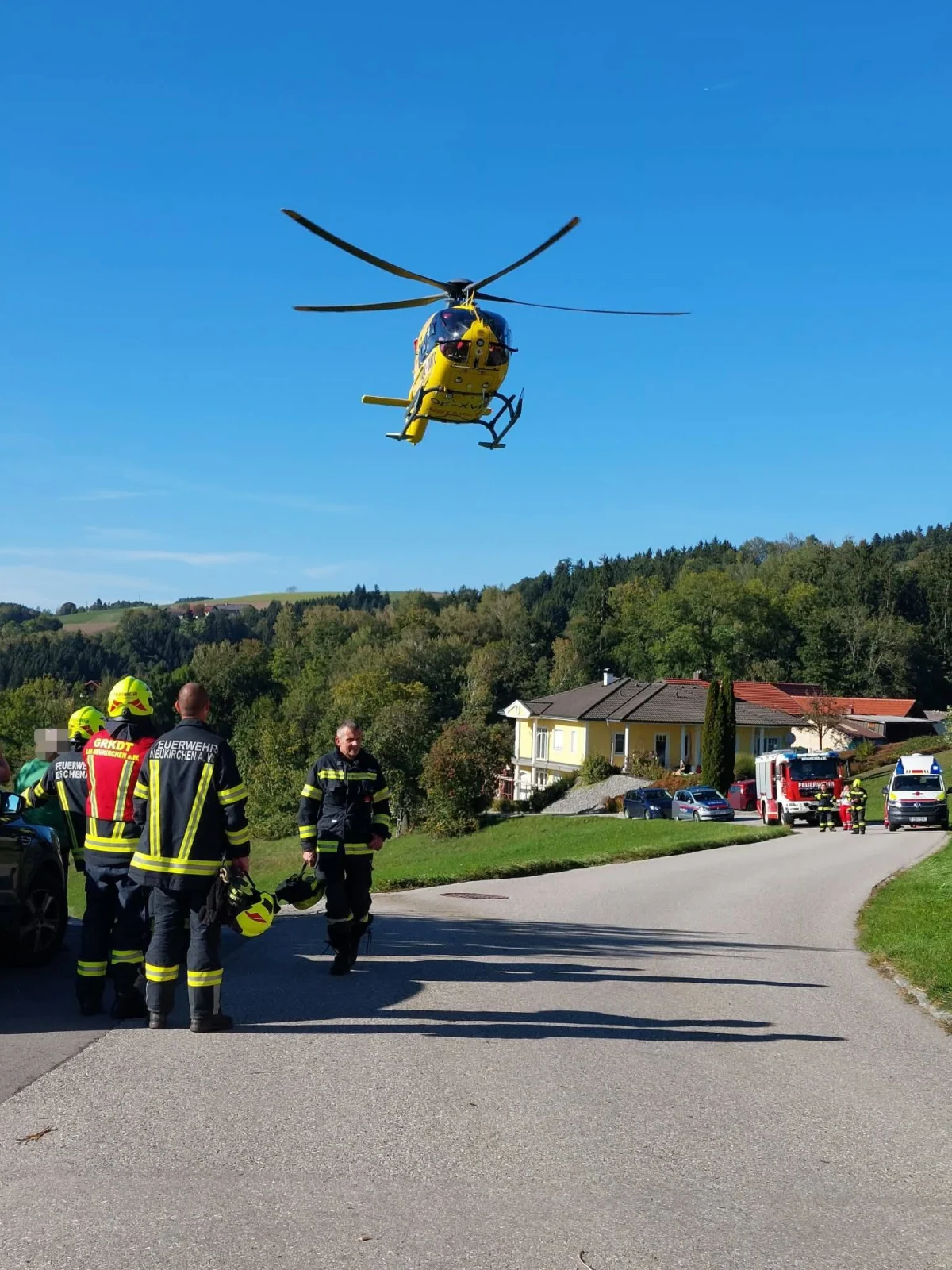 Mehrere Feuerwehrleute stehen auf einer ländlichen Straße, während ein gelber Rettungshubschrauber darüber schwebt. Im Hintergrund sind Wohnhäuser und Einsatzfahrzeuge sichtbar.