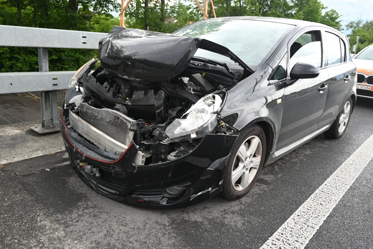 Ein schwarzes Auto mit erheblichen Schäden an der Front steht auf einer Straße nahe einer Leitplanke. Ein weiteres Fahrzeug ist im Hintergrund sichtbar.