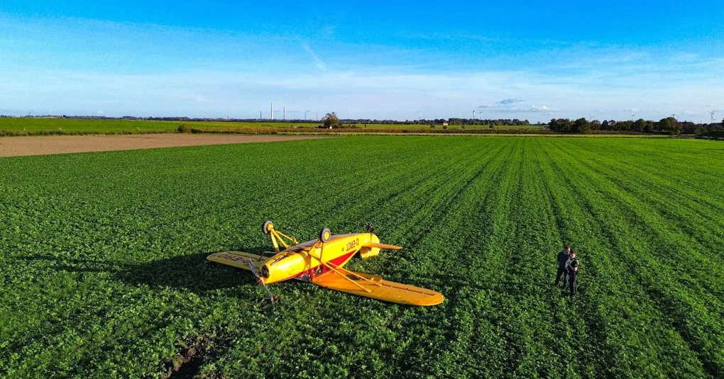 Ein gelbes Flugzeug ist kopfüber auf einem großen grünen Feld gelandet. Zwei Personen stehen daneben und begutachten den Schaden.