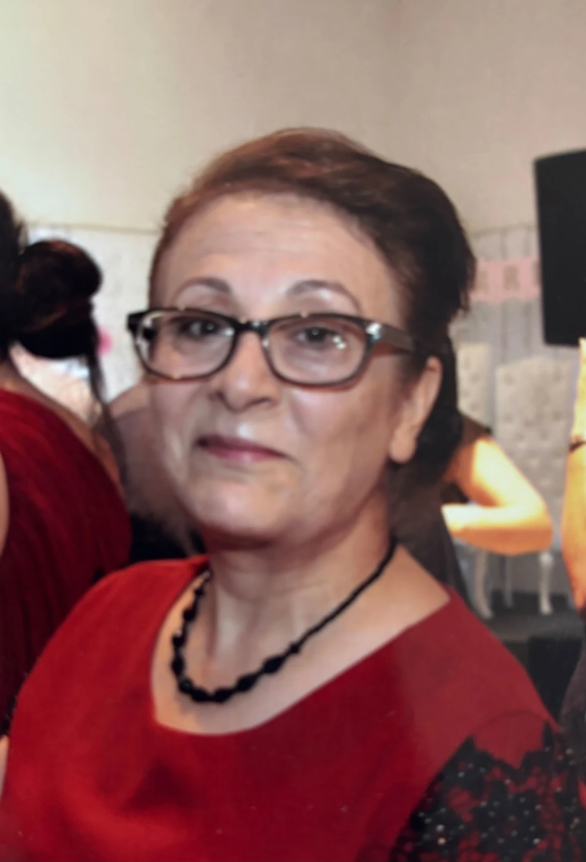 Eine Frau mittleren Alters trägt eine Brille und eine rote Bluse, während sie zu einer Kamera schaut.