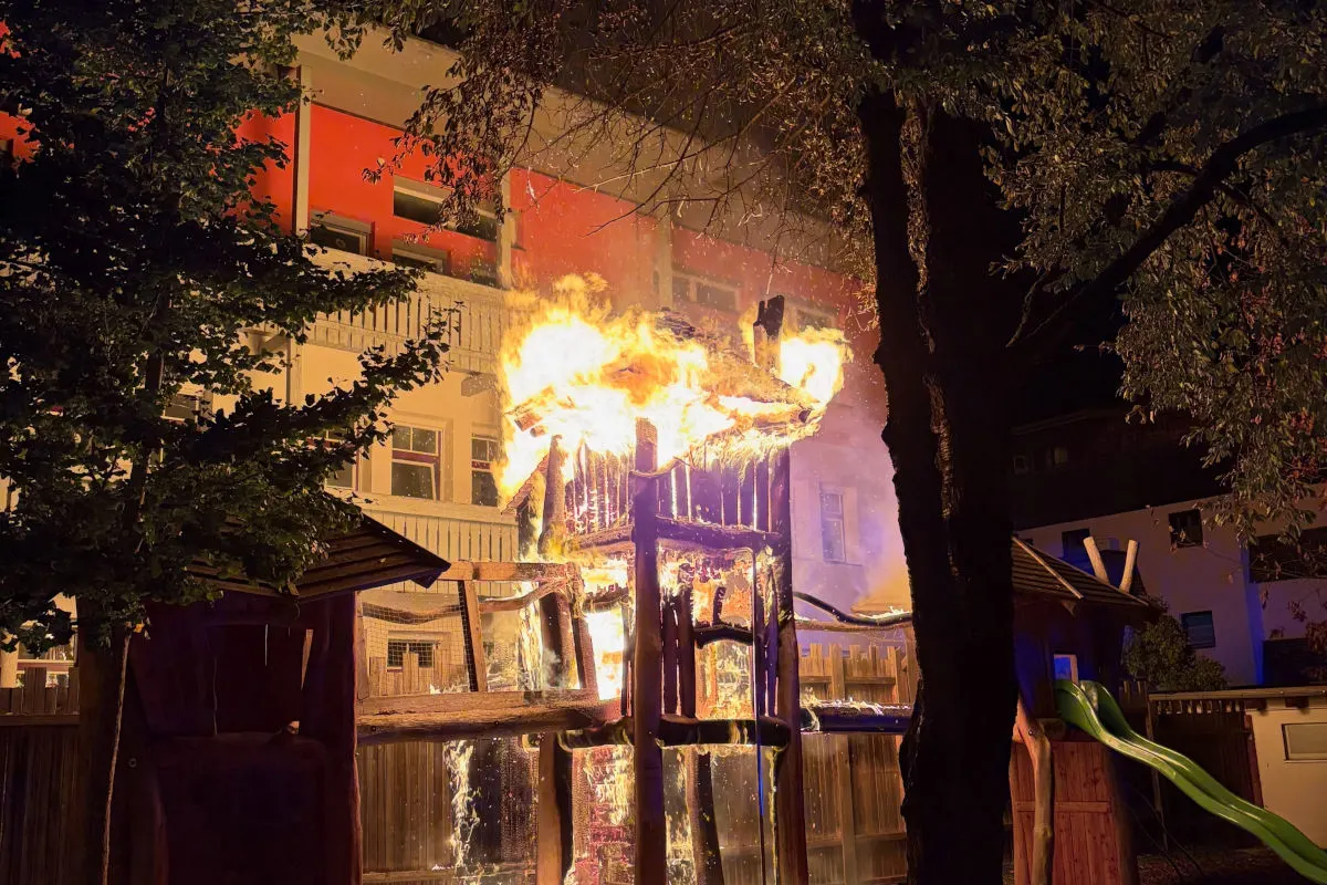 Ein Holzspielgerüst steht vor einem Wohngebäude in Flammen, umgeben von Bäumen, bei Nacht.