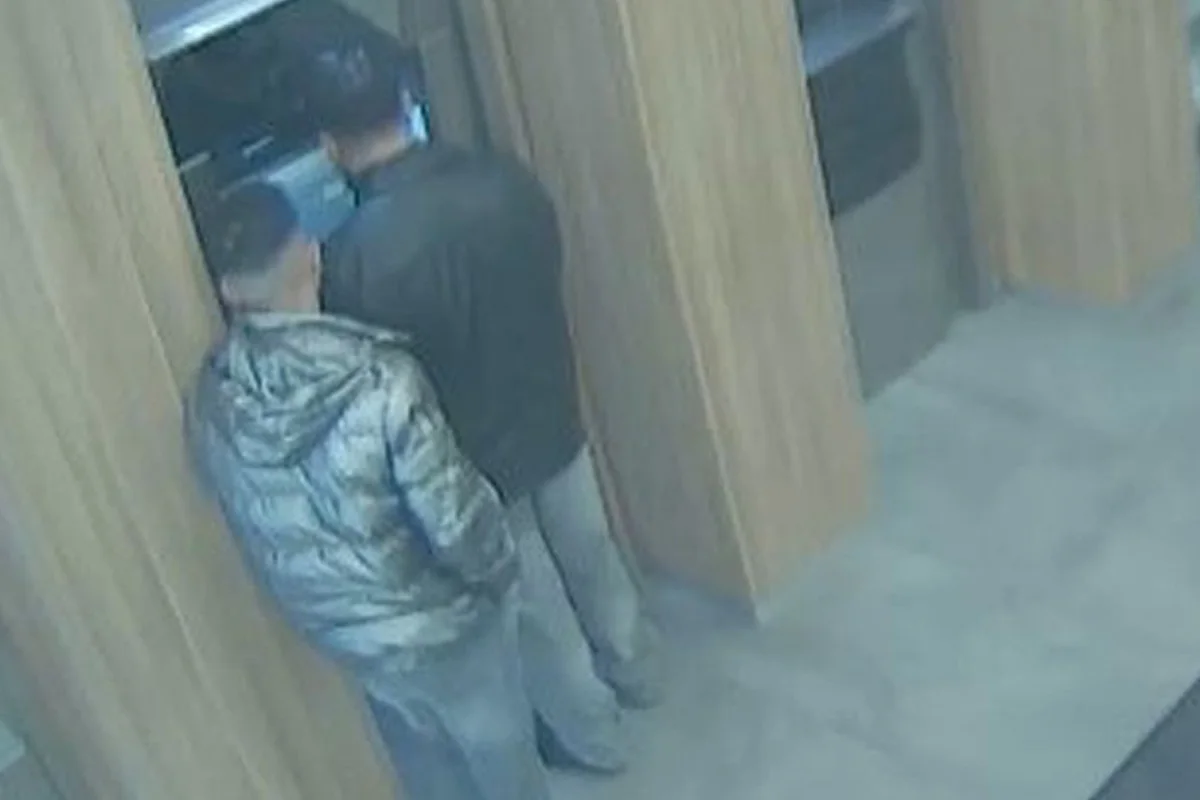 Zwei Personen stehen dicht nebeneinander in einem kleinen Raum vor einem Automaten. Die Szene scheint von einer Überwachungskamera aufgenommen worden zu sein.