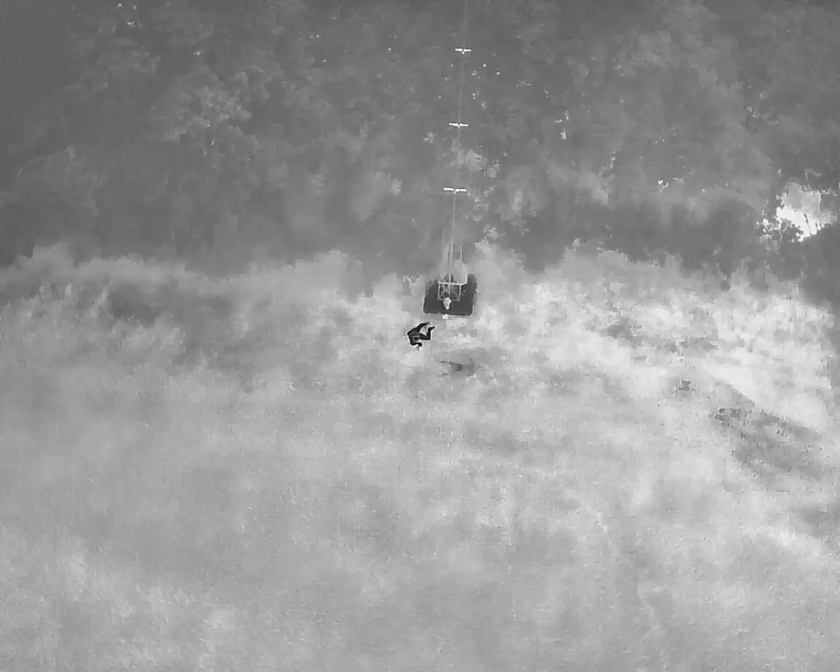 Eine Person in einem Geschirr wird von einem Hubschrauber mit einer Seilwinde aus einem Flussbett gehoben, umgeben von Bäumen.