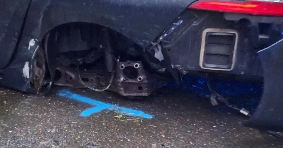 Ein beschädigtes schwarzes Auto zeigt ein fehlendes Hinterrad und hellblaue Markierungen auf dem Asphalt. Der Stoßfänger ist abgerissen.