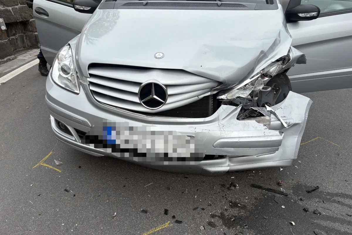 Ein silberner Mercedes, dessen rechte Vorderseite bei einem Unfall stark beschädigt wurde, steht auf einer Straße. Der Scheinwerfer ist zerbrochen und die Stoßstange ist eingedrückt.