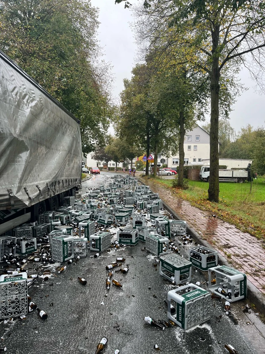 Auf einer nassen Straße liegen zahlreiche umgekippte Bierkisten und zerbrochene Glasflaschen verstreut. Bäume und Gehweg sind im Hintergrund sichtbar.