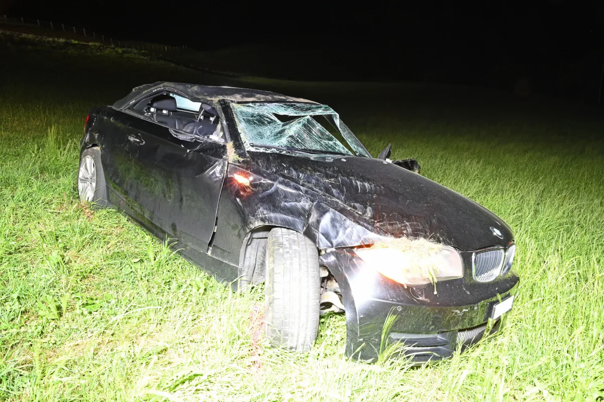 Ein stark beschädigtes schwarzes Auto steht nachts auf einer Wiese. Die Windschutzscheibe ist zerstört.